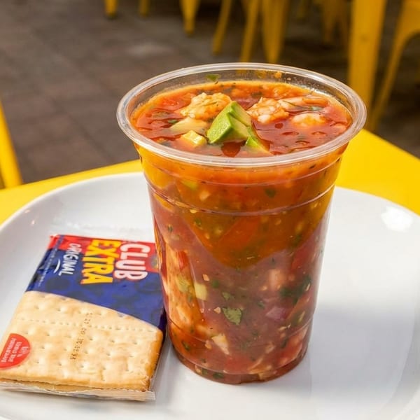 A plastic cup of ceviche with shrimp and avocado, alongside crackers on a white plate.