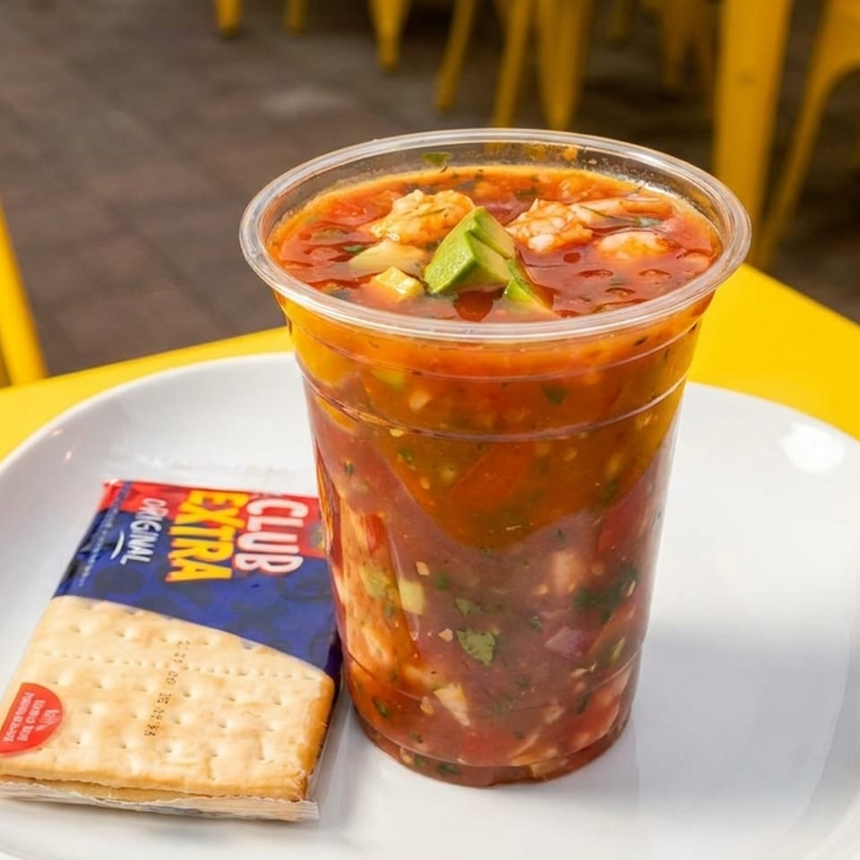 A plastic cup of ceviche with shrimp and avocado, alongside crackers on a white plate.